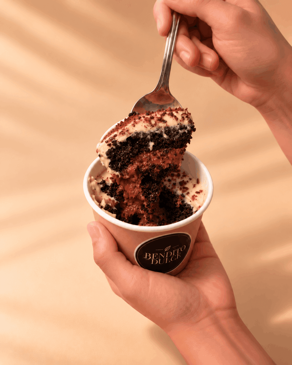Una mano sostiene un vasito de Bendito Dulce mientras la otra saca con la cuchara bizcochuelo de chocolate y crema